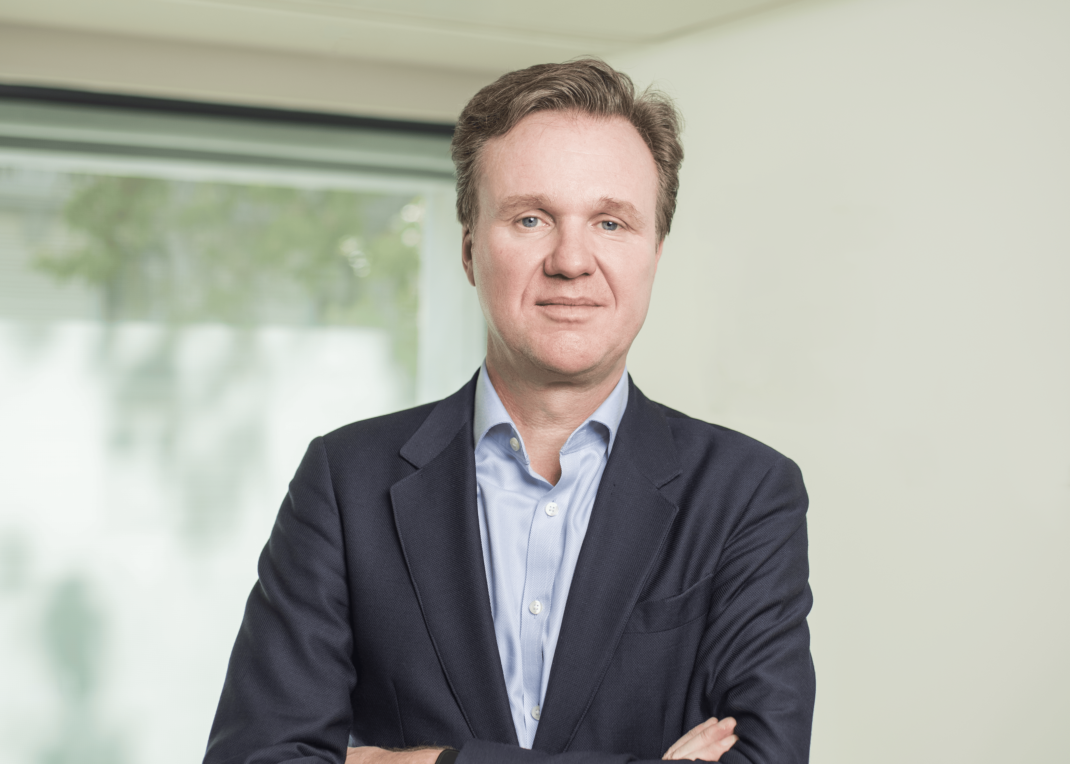 Professional portrait of a man with light brown hair in a navy blazer and light blue shirt, arms crossed, standing in a bright office.
