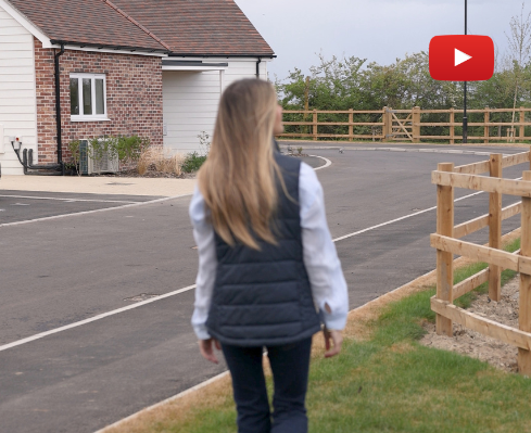 Person walking away from the camera along a residential street, with a brick house and wooden fence nearby; a YouTube play button overlay appears in the top-right corner.
